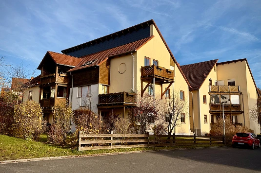 Mehrfamilienhaus mit gelb gestrichener Fassade, braunen Holzbalkonen und roten Ziegeldächern unter blauem Himmel.